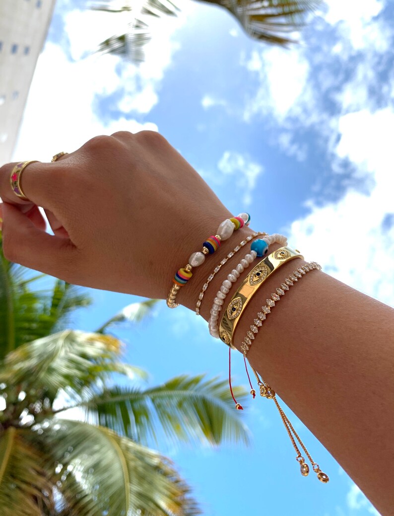 May include: A hand wearing five bracelets. The bracelets are a gold chain bracelet, a pearl bracelet, a gold bracelet with an evil eye design, a beaded bracelet with colorful beads, and a red string bracelet with gold beads.