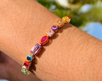 May include: A gold bracelet with colorful, rectangular and oval gemstones. The bracelet features red, purple, blue, green, and yellow stones. The bracelet is worn on a person's arm.