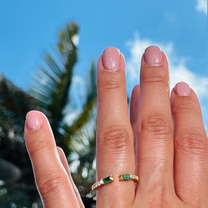 May include: A gold ring with a split design featuring two emerald green stones and a row of clear cubic zirconia stones.