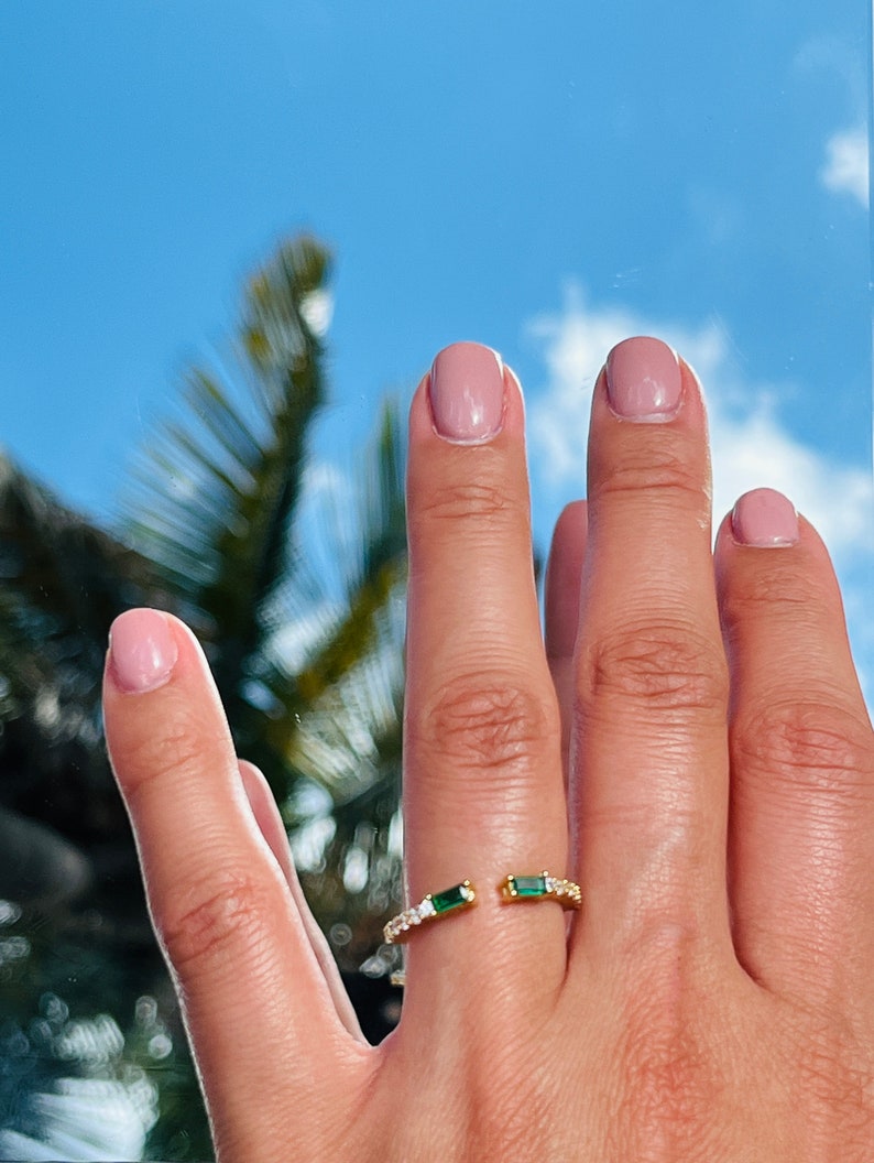 May include: A gold ring with a split design featuring two emerald green stones and a row of clear cubic zirconia stones.