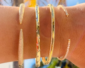 Puede incluir: Cuatro pulseras de oro con piedras transparentes. Las pulseras están apiladas en la muñeca de una persona.