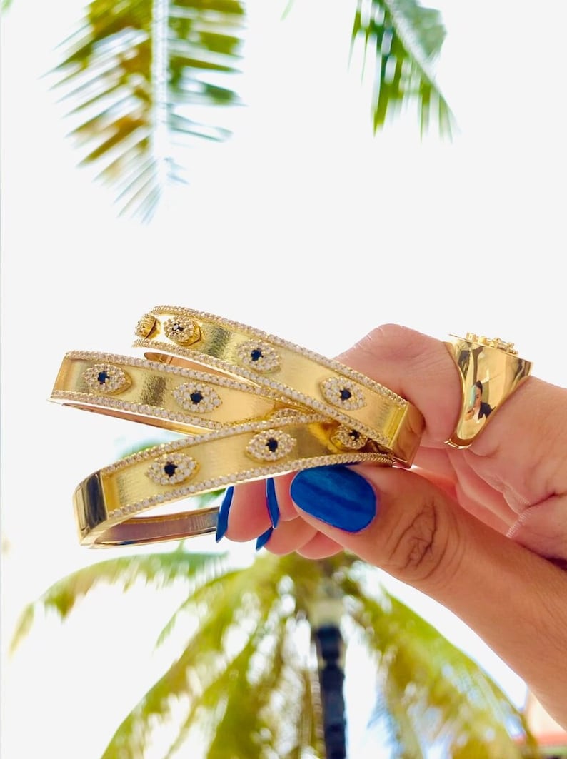 May include: Three gold bangles with a delicate design of small white stones and black evil eye charms. The bangles are stacked on top of each other and held in a hand with blue nail polish.