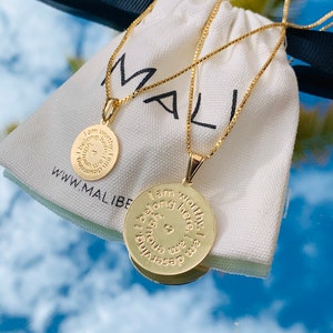Könnte beinhalten: Zwei goldene Halsketten mit runden Anhängern. Die Anhänger tragen die Worte "Ich bin würdig. Ich gehöre hierher. Ich bin genug. Ich bin es wert" eingraviert. Die Halsketten liegen auf einem weißen Stoffbeutel mit dem Wort "MALI" darauf.