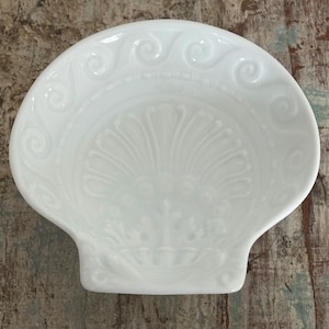 Puede incluir: Un plato de cerámica blanca con forma de concha de vieira. El interior presenta un diseño en relieve con un motivo central en forma de abanico que irradia desde la base, rodeado por un borde de patrones de espirales en el borde superior.