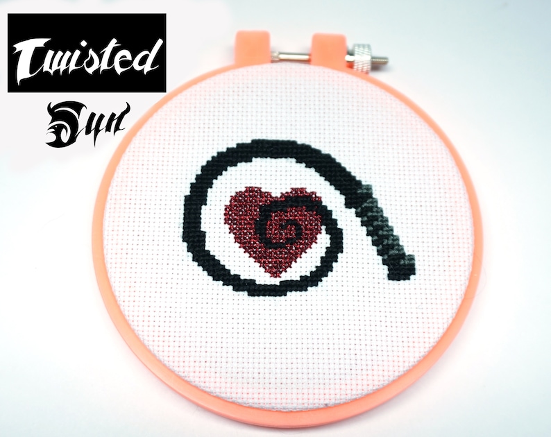 May include: A white embroidery hoop with a black and red cross-stitch design. The design features a heart with a spiral inside and a black whip wrapped around the heart.
