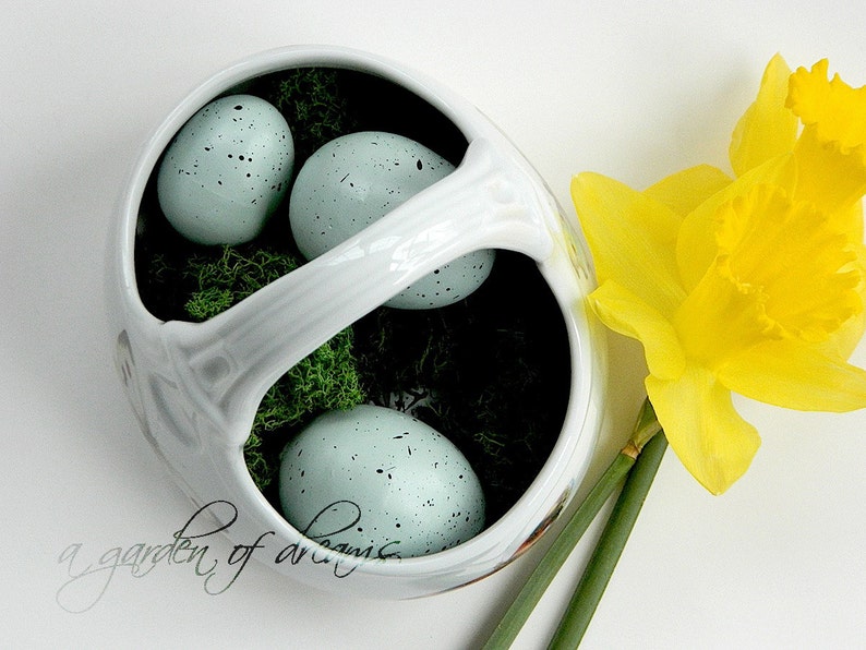 May include: A white ceramic basket with a handle, filled with green moss and four blue speckled eggs. The basket is sitting on a white surface next to a yellow daffodil.