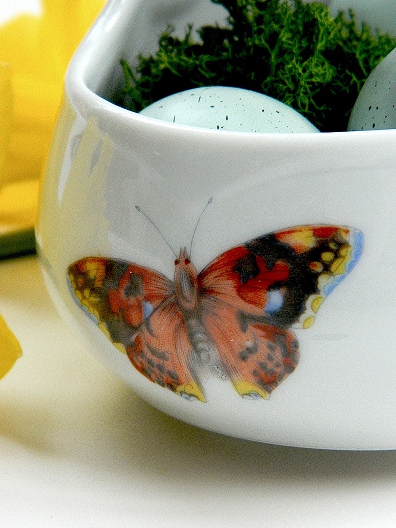 May include: A white ceramic bowl with a butterfly design. The bowl is filled with moss and blue speckled eggs.