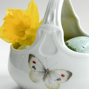 May include: A white ceramic basket with a handle, featuring a yellow daffodil and a butterfly with red, white and blue accents. The basket contains two speckled green eggs.