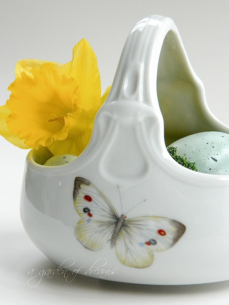 May include: A white ceramic basket with a handle, featuring a yellow daffodil and a butterfly with red, white and blue accents. The basket contains two speckled green eggs.