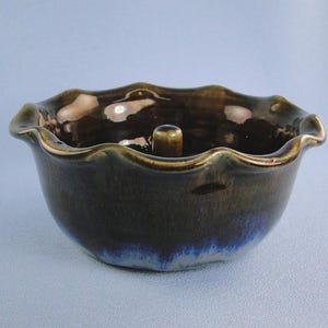 May include: A ceramic apple baker with a dark brown and blue glaze. The bowl has a wavy rim and a central stem holder. The exterior has a dark brown color, with blue hues at the base. The interior is glossy.