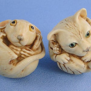 May include: Two small, cream-colored animal figurines. One is a mouse, the other a cat, both curled up. The mouse has small ears and a long tail. The cat has green eyes and detailed fur markings. They are on a blue surface.