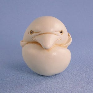 May include: A carved, off-white bird figurine with a stern expression. The bird has a prominent beak and small, dark eyes. The figurine is egg-shaped, with a rounded base and a detailed head. The background is a solid light blue.