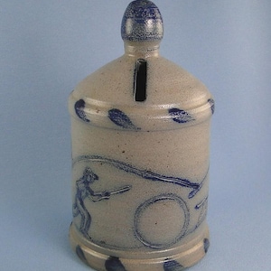May include: A stoneware bank with a cylindrical body and a rounded top. The bank is beige with blue decorative accents and a blue illustration of a person playing a sport. A slot is cut into the top for inserting coins.