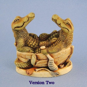May include: A detailed figurine featuring two alligator-like creatures in a small boat. The figures are tan and green, with textured skin and expressive faces. The boat is made of wood-like material, and the base is beige. The text "Version Two" is at the bottom.