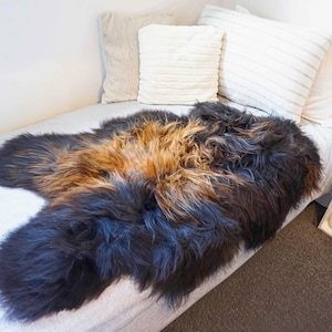 May include: A sheepskin rug in shades of brown, black, and tan lies on a bed. The rug's fluffy texture contrasts with the bed's white linen. Pillows in cream and white are behind the rug. A small white shelf is visible.