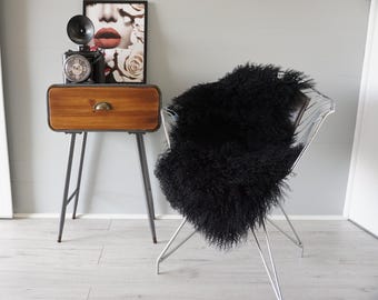 May include: A black, long-haired sheepskin rug draped over a wire-frame chair with thin metal legs. The rug has a dense, curly texture and covers the seat and backrest of the chair, providing a soft contrast to the structured metal frame.