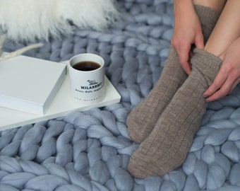 May include: A person wearing brown socks sits on a chunky knit blue blanket with a white mug of coffee and a white notebook on a white surface.