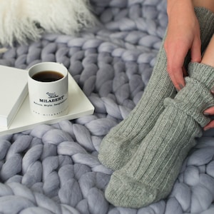 May include: A close-up of a person's feet wearing grey knitted socks. The person is sitting on a large, chunky knit blanket. A white mug with the text "MILABERT" is on a white surface in the background.
