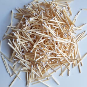Puede incluir: Un montón de cerillas de madera con puntas blancas. Las cerillas están dispersas, creando un patrón texturizado. La madera tiene un color marrón claro natural.