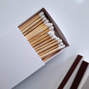 Może przedstawiać: Białe pudełko wypełnione drewnianymi zapałkami z białymi główkami. Pudełko jest otwarte, a zapałki są widoczne.