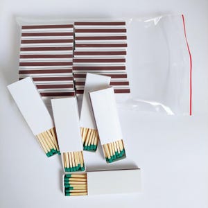 Pode incluir: Um close-up de várias caixas de fósforos com pontas verdes. As caixas são brancas e têm uma faixa marrom na lateral. Os fósforos estão dispostos em uma pilha sobre uma superfície branca.