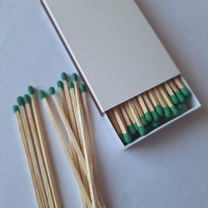 Peut inclure: Une boîte d'allumettes blanche, partiellement ouverte, révélant des allumettes en bois avec des têtes vertes. Plusieurs allumettes sont éparpillées à côté de la boîte. Les allumettes sont de couleur bois naturel.
