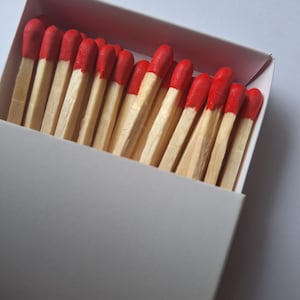 May include: A close-up of a matchbox filled with matches. The matches have red tips and light brown wooden stems. The matchbox is white with a red interior.