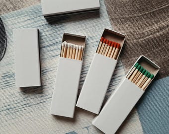 May include: Four rectangular matchboxes with plain white covers. One box is closed, while three are partially open, revealing wooden matches with white, red, and green tips respectively. The matchboxes are arranged in a cluster on a flat surface.