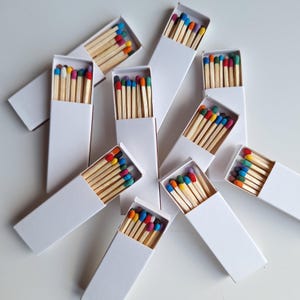 Puede incluir: Varias cajas de cerillas blancas están dispersas sobre una superficie blanca. Cada caja contiene cerillas de madera con puntas de colores en varios tonos, incluyendo rojo, azul, amarillo y verde. Las cerillas están ordenadas dentro de las cajas.