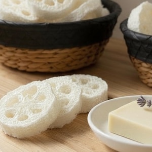 May include: A spa-themed image featuring natural bath products. Includes white loofah sponges, a bar of soap, and decorative lavender sprigs. The loofahs are in a black and woven basket. The soap is in a small white dish. The scene is set on a wooden surface.