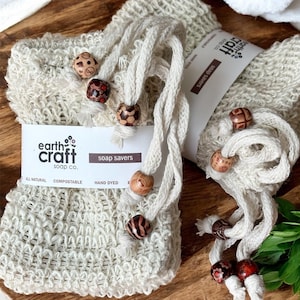 May include: Two beige soap savers made of woven material with cream-colored rope handles and decorative wooden beads. The label reads "earth craft soap co." and "soap savers."