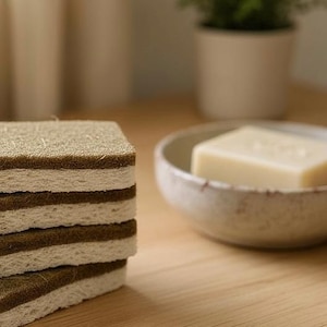 May include: A stack of square, eco-friendly cleaning sponges with a textured, brown top layer and a white, absorbent base. A bar of soap sits in a small, ceramic bowl. The scene is set on a wooden surface, suggesting a focus on natural home goods.