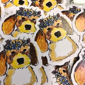 Op de afbeelding: Een verzameling stickers met een cartoonhond met bruine en gele vacht, met een blauwe bloemenkrans. De hond heeft een zwarte neus en ogen. De stickers staan op een witte achtergrond.