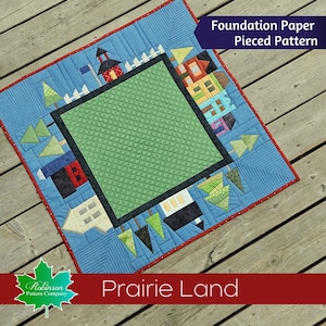 以下が含まれることがあります： 「プレーリーランド」というタイトルのキルトのためのファウンデーションペーパーピースキルトパターン。キルトは、中央に緑色の正方形があり、その周りを青い縁取りで囲み、家、木、フェンスが描かれています。キルトは木製の表面の上に置かれています。