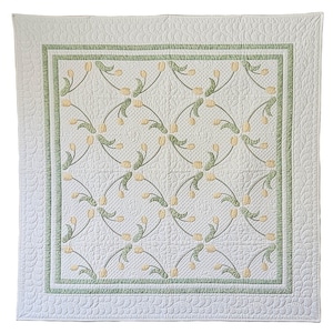 May include: A white quilt with a repeating pattern of yellow tulip-like flowers and green stems. The quilt has a green border and a scalloped edge. The quilting creates a textured effect on the white fabric.