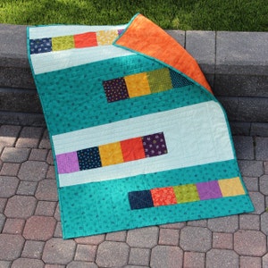 Puede incluir: Una colcha de color turquesa y blanco con un diseño de patchwork que presenta cuadrados de varios colores, incluyendo naranja, amarillo, morado y verde.