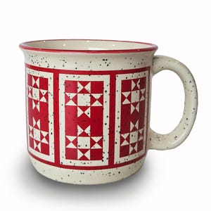 May include: Cream-colored ceramic mug with a red and white star quilt pattern. The mug has a speckled finish and a rounded handle. The rim and the inside of the mug are red.