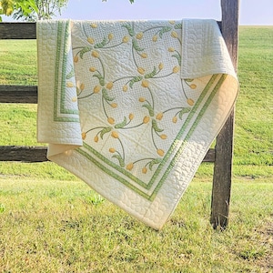 May include: A cream-colored quilt with a floral pattern of yellow tulip-like flowers and green stems. The quilt has a green border and is draped over a wooden fence in a grassy field. The quilt is a handmade item.