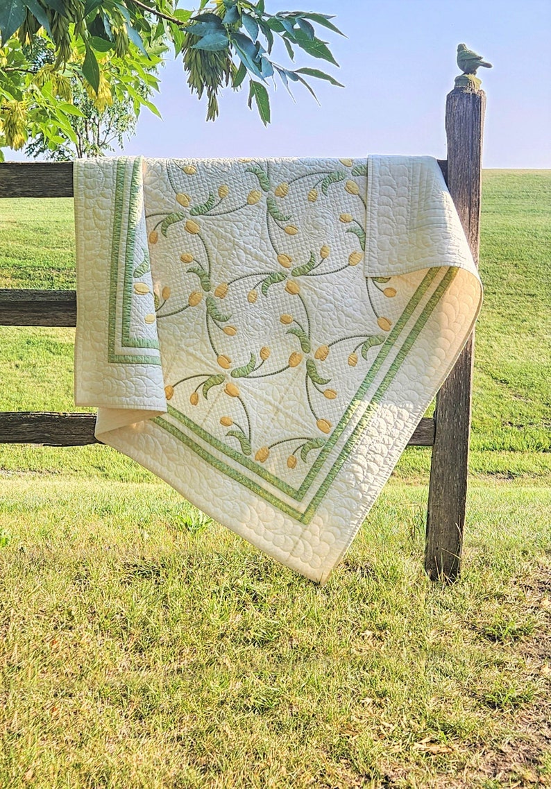 May include: A cream-colored quilt with a floral pattern of yellow tulip-like flowers and green stems. The quilt has a green border and is draped over a wooden fence in a grassy field. The quilt is a handmade item.