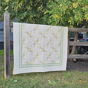 May include: A cream-colored quilt with a repeating pattern of yellow flowers and green stems. The quilt has a green border and is hanging on a wooden fence. The quilt is displayed outdoors in a grassy area.