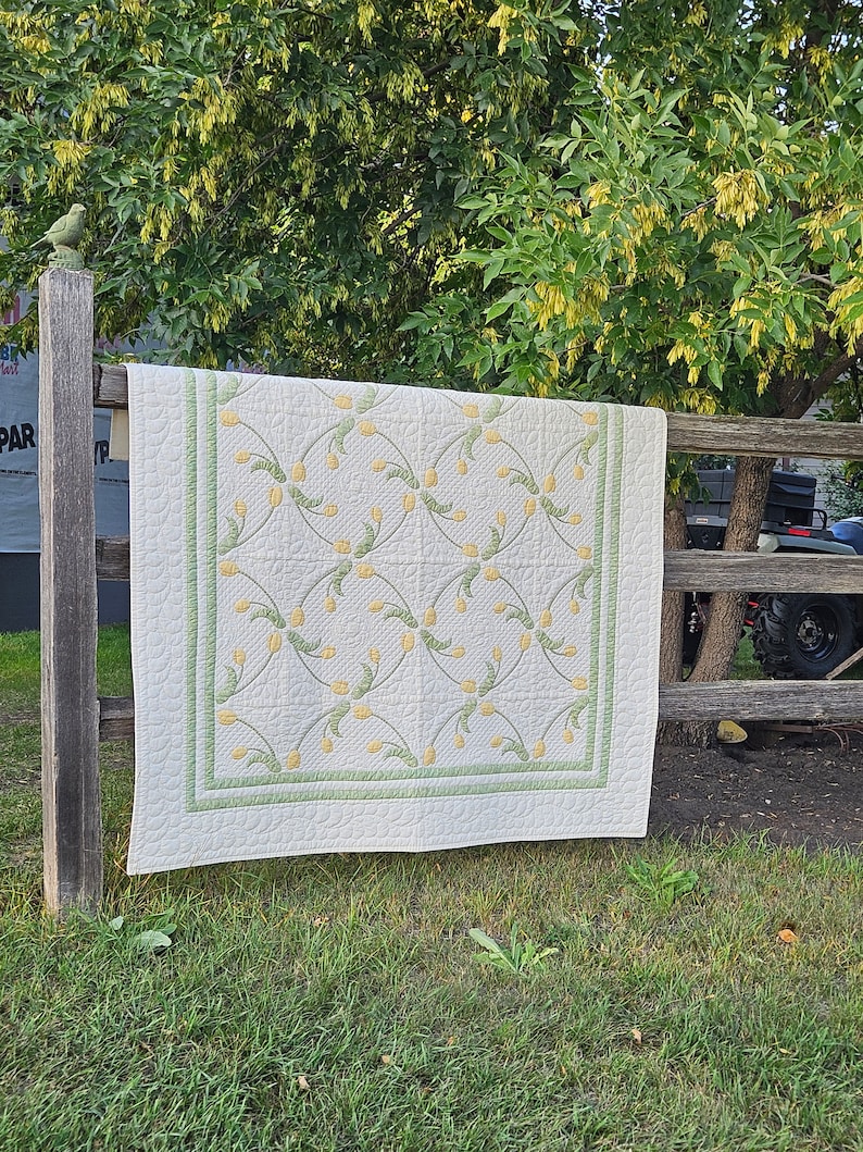 May include: A cream-colored quilt with a repeating pattern of yellow flowers and green stems. The quilt has a green border and is hanging on a wooden fence. The quilt is displayed outdoors in a grassy area.