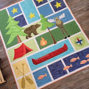 Op de afbeelding: Een kleurrijk quilt met een kampeerthema met een rode tent, een kano, een beer, een eland, een kampvuur en een kompas. De quilt is gemaakt van verschillende vierkanten en rechthoeken met verschillende kleuren en patronen.