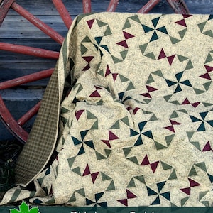 Op de afbeelding: Een beige, groene en bordeauxrode quilt met een geometrisch patroon van windmolens. De quilt is gevouwen en gedrapeerd over een houten wagenwiel. De tekst "Oklahoma Twister" is op de quilt gedrukt.