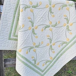 May include: A cream-colored quilt with a floral design featuring yellow tulip-like flowers and green stems. The quilt has a green border and a scalloped edge. The quilt is draped over a wooden fence.