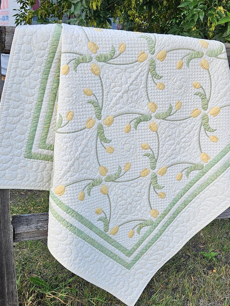 May include: A cream-colored quilt with a floral design featuring yellow tulip-like flowers and green stems. The quilt has a green border and a scalloped edge. The quilt is draped over a wooden fence.