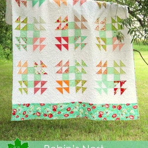 May include: A white quilt with a "Robin's Nest" pattern featuring green, pink, and orange triangles. The quilt is hanging on a tree branch and has a green and white floral border.