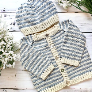 May include: A baby's knitted set, including a striped cardigan and matching beanie. The set features alternating light blue and cream-colored horizontal stripes. The cardigan has button closures and the set is displayed on a white wooden surface.