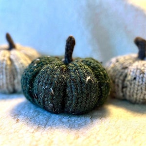 May include: Three knitted pumpkins in shades of green, white, and gray. Each pumpkin has a brown stem. The pumpkins are arranged on a soft, white surface, creating a cozy, autumnal theme.