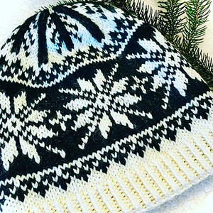 May include: A knitted winter hat with a black and white snowflake pattern. The hat has a ribbed cream-colored brim and a black and white design on the crown. Green pine needles are in the background.
