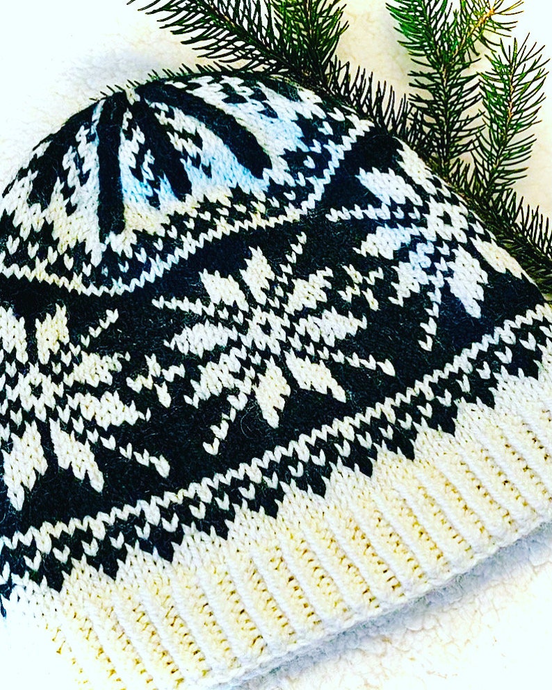 May include: A knitted winter hat with a black and white snowflake pattern. The hat has a ribbed cream-colored brim and a black and white design on the crown. Green pine needles are in the background.